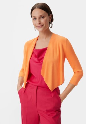 Cardigan arancione lavorato a maglia con un davanti drappeggiato e maniche a tre quarti, abbinato a una blusa in satin rosa e pantaloni rosa sartoriali.