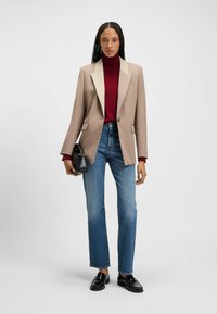 Blazer beige con colletto bianco a contrasto, abbinato a un dolcevita bordeaux, jeans blu a gamba dritta e mocassini neri.