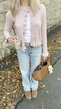 Cardigan a maglia rosa chiaro con bordi arricciati, indossato sopra una maglietta bianca. Jeans azzurri, stivali color cammello e una borsa marrone con un accento in peluche.