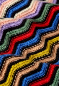 Wellig gestricktes Muster in verschiedenen Farben, darunter Rot, Blau, Schwarz, Grün und Gelb. Sanfte Textur mit einem ausgeprägten Zickzackdesign und kontrastierenden Streifen.