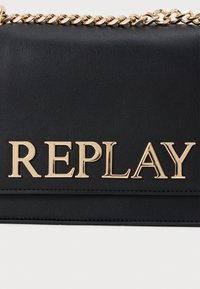 Čierna kabelka z umelej kože s pozláteným reťazovým popruhom a vytlačeným logom "REPLAY" zlatými blokovými písmenami na prednej strane.