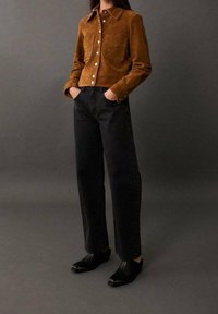 Nainen, jolla on yllä ruskea nappikiinnitteinen nahkatakki, mustat korkeavyötäröiset leveälahkeiset farkut ja mustat neliönmalliset nilkkasaappaat, kädet taskuissa, harmaa tausta.