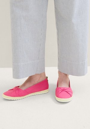 Voeten met felle roze instapschoenen met een knoopontwerp, gecombineerd met lichtgrijze broek met wijde pijpen tegen een neutrale achtergrond.