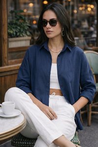 Vrouw in marineblauw shirt en witte broek met een zonnebril, zittend buiten aan een cafétafel met een wit koffiekopje.