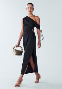 Robe noire à une épaule en matière lisse, présentant une fente sur le côté, des détails froncés et une découpe à la taille. Accessoirisée avec un sac tissé doré.