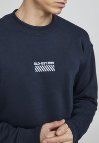 Sudadera azul marino con cuello redondo acanalado y un pequeño diseño bordado en blanco con rayas y el texto "SLD-EST:1999" en el pecho.