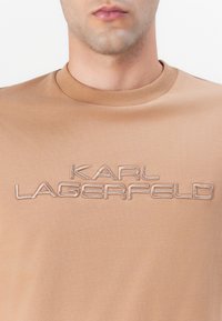 Smėlio spalvos medvilninė marškinė su apvalia iškirpte, pasižyminti šviesiai ruda siuvinėta užrašu "KARL LAGERFELD" per krūtinę. Lygi tekstūra.