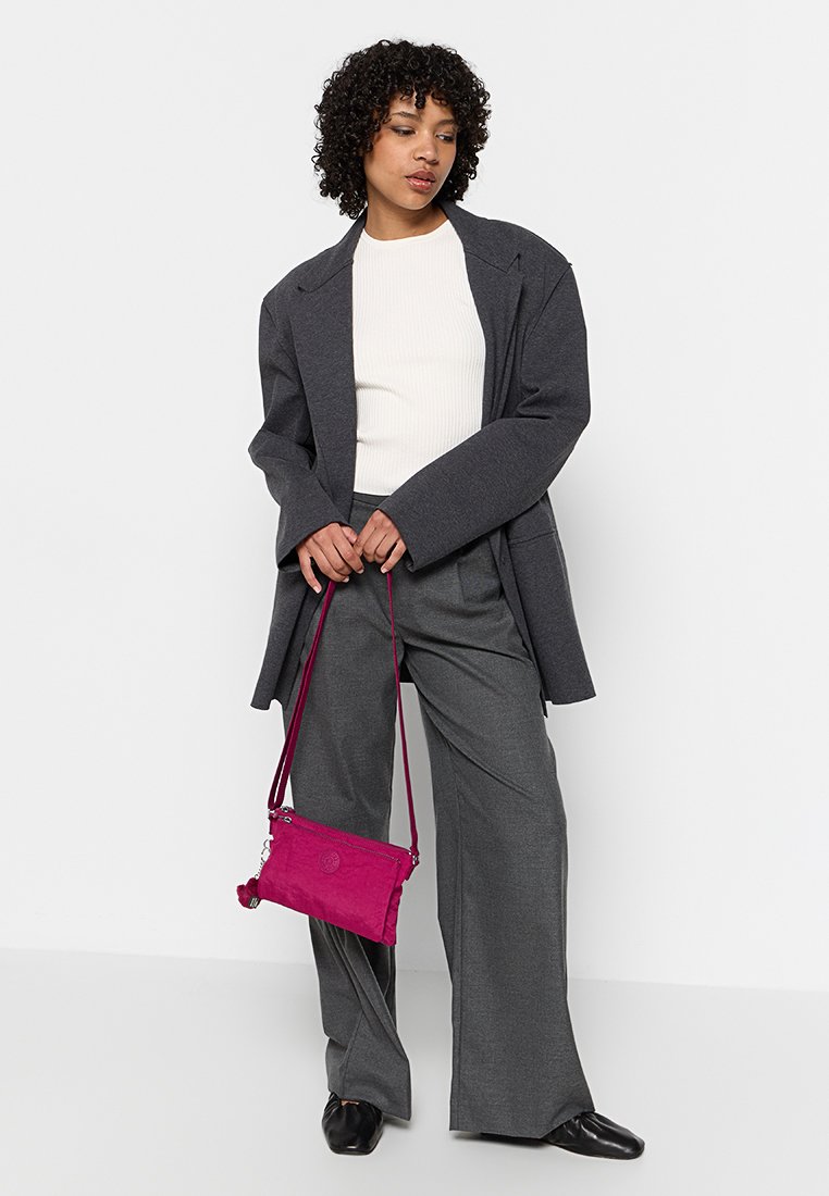 Donna con giacca grigia scura e pantaloni a gamba larga, top bianco, che tiene una piccola borsa a tracolla rosa brillante su uno sfondo bianco.