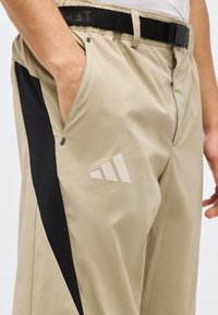 Μπεζ παντελόνι με μαύρη πλευρική ρίγα και ενσωματωμένη μαύρη ζώνη, που φοριέται από άτομο με το αριστερό χέρι στην τσέπη, με ανακλαστικό τριγωνικό λογότυπο.