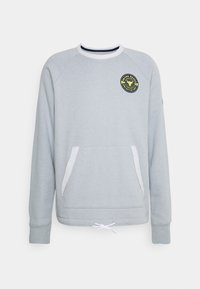 Sudadera gris claro con cuello redondo, mangas raglán, bolsillo delantero y un parche redondo negro y amarillo en el pecho. Cordonera en el dobladillo.