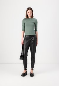 Ljust grön figurnära långärmad topp, kombinerad med svarta läderbyxor i tapered-modell. Modellen håller en liten svart handväska och bär kraftiga svarta skor.