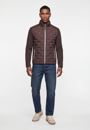 Mann trägt eine braune gesteppte Jacke mit Reißverschluss, blaue Jeans und beige Sneakers und steht mit entspannten Händen an den Seiten vor einem weißen Hintergrund.