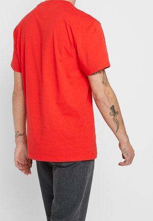 Man wearing a plain red short-sleeve T-shirt and dark gray jeans, showing tattoos on both forearms and upper arm against a white background.