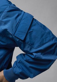 Manga de chaqueta de nailon azul con un bolsillo con solapa y puño acanalado contra un fondo gris.