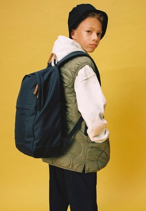 Zaino blu navy con dettagli beige, dotato di una tasca frontale e spallacci imbottiti. Indossato sopra un gilet trapuntato verde e una felpa bianca con cappuccio.