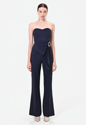 Femme portant une combinaison bleu marine sans bretelles avec un détail de boucle sur le côté et un pantalon à jambes larges, associée à des talons pointus argentés et de longues boucles d'oreilles.