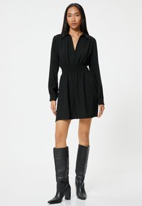 Schwarzes Hemdkleid mit einem Kragen, geraffter Taille, langen Ärmeln und einem ausgestellten Rock. Kombiniert mit kniehohen schwarzen Lederstiefeln.