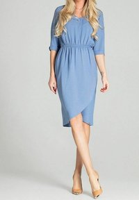 Frau in einem hellblauen Kleid mit langen Ärmeln bis zum Ellenbogen und tailliertem Schnitt, die selbstbewusst steht und nude-farbene High Heels trägt, vor einem schlichten Hintergrund.