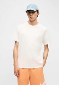 Mladý muž nosiaci svetlomodrú čiapku, jednoduché biele tričko a oranžové šortky s bielym logom, stojaci s jednou rukou vo vrecku, neutrálne pozadie.