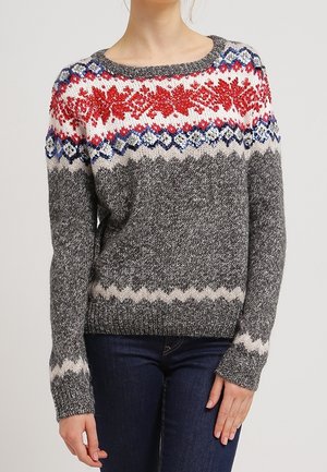 Femme portant un pull en maille avec des motifs nordiques rouges, blancs, bleus et gris sur la poitrine et le haut des manches, associé à un jean bleu foncé.