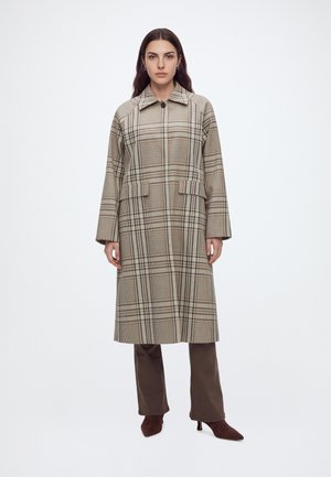 Femme portant un long manteau beige à carreaux par-dessus un pantalon marron et des chaussures marron à talons, debout devant un fond blanc uni.