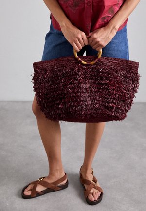 Burgundy színű, szőtt vászontáska, frizurás textúrával, kerek fa fogantyúkkal és széles, nyitott tetővel. Farmer rövidnadrággal és barna szandálokkal párosítva.