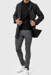 Grauer Wollmantel über einem schwarzen, strukturierten Pullover, kombiniert mit grauen Jeans mit gerippten Akzenten und weißen Turnschuhen.