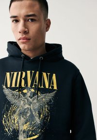 Melna kapuce ar zelta "NIRVANA" drukājumu un eņģeļa grafiku. Ietver auklas un mīkstu tekstūru. Standarta piegriezums.