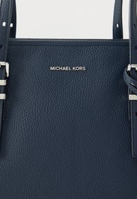 Merensininen nahkainen käsilaukku, jossa on teksturoitu pinta, hopeiset metalliosat ja etuosassa kohopainettu "MICHAEL KORS" -logo.