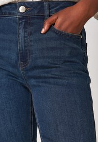 Mano con tonalità di pelle scura che riposa all'interno della tasca anteriore di jeans blu in denim con cuciture visibili e bottone di metallo.