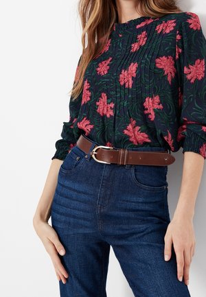 Femme portant un chemisier floral sombre avec des fleurs roses, rentré dans un jean bleu taille haute maintenu par une ceinture en cuir marron avec une boucle argentée.