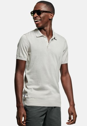 Lachende man met donkere zonnebril, lichtgrijs polo met korte mouwen en donkere broek, kijkend naar links tegen een effen achtergrond.