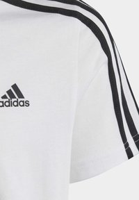 Camiseta blanca de manga corta con el logo negro de Adidas y tres franjas negras a lo largo de los hombros. Material de algodón suave, diseño de cuello redondo.