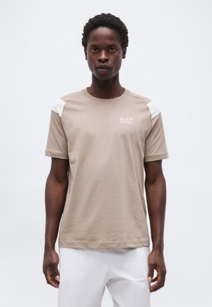 Άνδρας που φοράει μπεζ μπλουζάκι EA7 Emporio Armani με λευκές λεπτομέρειες στους ώμους, στέκεται μπροστά σε απλό ανοιχτόχρωμο φόντο.