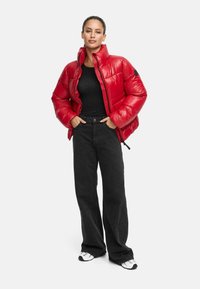 Giacca puffer rossa con colletto alto e chiusura a zip, caratterizzata da una texture lucida. Abbinata a pantaloni neri a gamba larga e sneakers bianche.