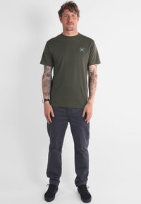 Camiseta verde oliva con cuello redondo y mangas cortas; combinada con pantalones gris oscuro. Zapatos negros y tatuajes visibles en ambos brazos.