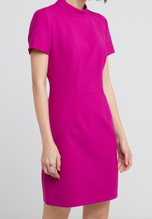 Femme portant une robe ajustée à manches courtes de couleur rose vif, avec des coutures décoratives et un col montant, debout devant un fond clair uni.