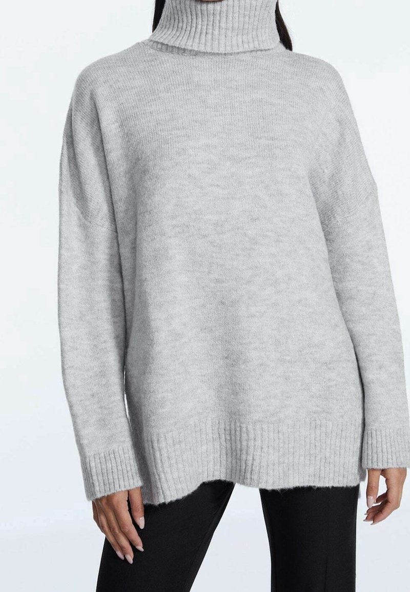 Femme portant un pull à col roulé gris clair oversize avec des poignets et un ourlet côtelés, associé à un pantalon noir.
