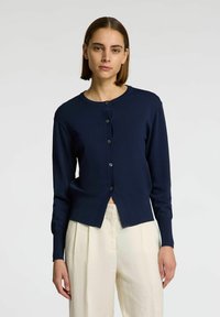 Námorně modrý cardigan s dlouhými rukávy, kulatým výstřihem, předními knoflíky a mírně zkráceným designem. Hladká pletená textura a minimální detaily.