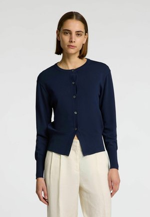 Námorně modrý cardigan s dlouhými rukávy, kulatým výstřihem, předními knoflíky a mírně zkráceným designem. Hladká pletená textura a minimální detaily.