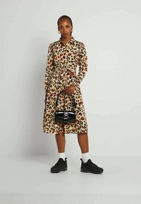 Mulher em pé vestindo um vestido de estampa de leopardo com cinto, meias brancas, tênis pretos, segurando uma bolsa preta com detalhe de fivela metálica.