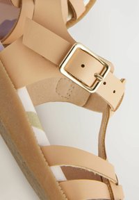 Beige lädersandal med ett remdesign, med en detalj i guldspänne. Texturerad innersula med ett blommönster. Platt sula.
