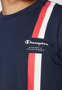 Camiseta de algodón azul marino con llamativas rayas verticales rojas y blancas, y un logo de Champion centrado con el texto "AUTHENTIC ATHLETICWEAR."