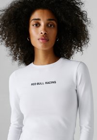 Kobieta z kręconymi włosami, ubrana w białą koszulę z długim rękawem z napisem "RED BULL RACING" na piersi, neutralne szare tło.