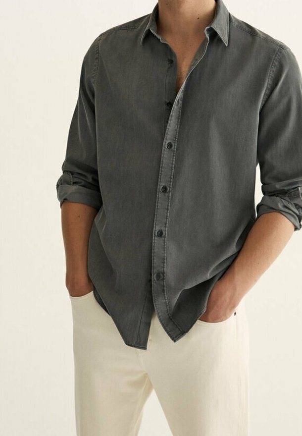 Homme portant une chemise gris foncé à boutons avec manches retroussées et un pantalon beige clair, les mains dans les poches, debout devant un fond uni.