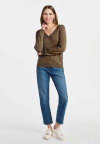 Brązowy sweter z dekoltem w serek, o miękkiej fakturze, luźny krój, w zestawieniu z niebieskimi dżinsami i białymi trampkami. Modelka pozuje z ręką na brodzie, uśmiechając się.