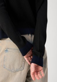 Une personne se tient dos à l'appareil photo, portant un pull noir avec des poignets bleu marine et un jean en denim clair, les mains jointes derrière.