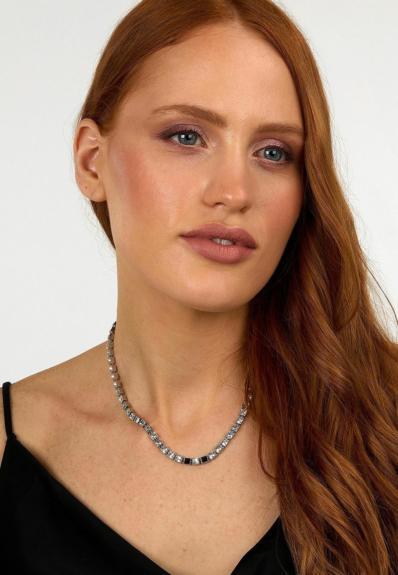 Femme aux longs cheveux roux portant un haut noir et un collier argenté avec des maillons carrés sur un fond uni.