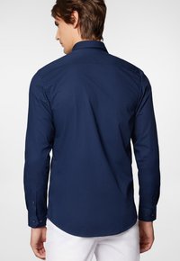 Granatowa koszula z długim rękawem, dopasowany krój, detal wzmocnienia na plecach i zaokrąglone wykończenie. Tkanina wydaje się gładka o matowym wykończeniu.
