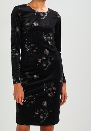 Czarna aksamitna sukienka za kolano z długimi rękawami i subtelnym wzorem dmuchawca, noszona przez stojącą osobę.